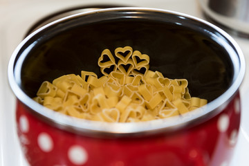 Heart shaped pasta in red pot with white dots on stove. Concept: preparing meal with love

