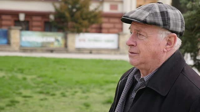 Portrait Of An Optimistic Old Man In A Checkered Cap And A Raincoat Walking In A Park Lane On A Sunny Day In Spring In Slow Motion