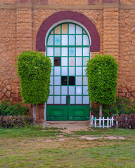 Closed grunge door with green metal grid framed by two green bushes in orange colored bricks stone wall in sunrise time at Montaza public park, Alexandria, Egypt