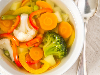 Bright spring vegetable soup with cauliflower, broccoli, pepper, carrot, green peas. Top view, white wooden background.
