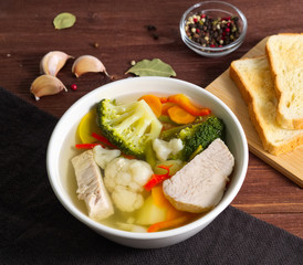Bright dietary soup with a chiken meat, cauliflower, broccoli and other vegetable on brown wooden background, side view.
