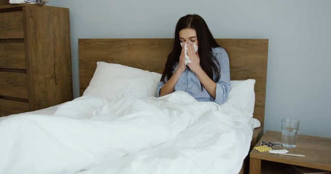 Unhealthy young brunette woman sitting in the bed, sneezing in the napkin and feeling herself bad as having a cold. Indoors