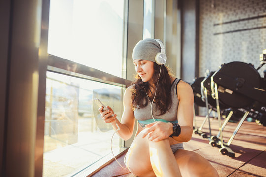 Theme sports, health and technology. beautiful sexy Caucasian woman sportswoman in gray sportswear and hat sits by window with sun setting uses smartphone listen music in large headphones on head