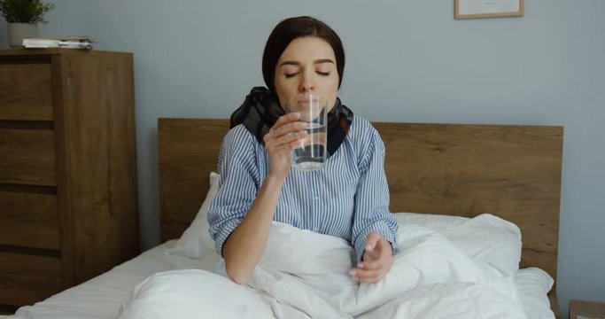 Unwell young Caucasian woman in a scarf and pajama sitting in the bed and taking a pill with water as having a temperature. Indoor