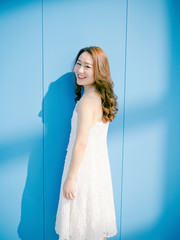 Beautiful Chinese brunette woman in white wedding dress posing and smiling at camera against blue wall background. Sunny portrait of glamour young stylish lady. Emotions, beauty and lifestyle concept.