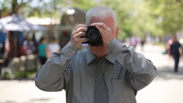 A cheery view of a grey-headed man with a short haircut in a grey shirt and a necktie standing in a street and taking photos in summer 