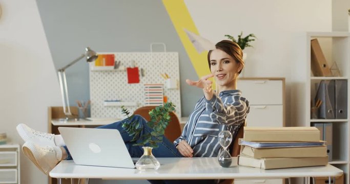 Young beautiful woman sitting at the office table, holding her legs on it, making a paper plane and throwing it away. Indoor