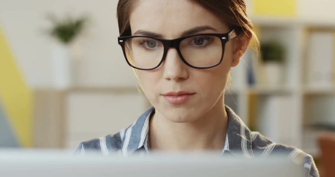Portrait of the young attractive serious woman in the striped shirt and glasses working in front of the computer screen in the office. Inside