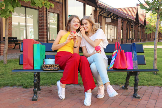 Beautiful women with shopping bags and cups of coffee sitting on bench outdoors