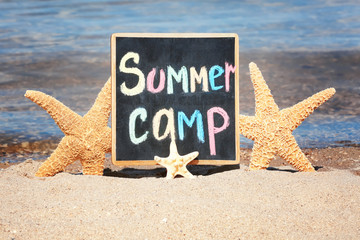 Little blackboard with text SUMMER CAMP and sea stars on sand near sea
