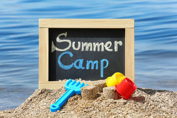 Little blackboard with text SUMMER CAMP and toys on sand near sea