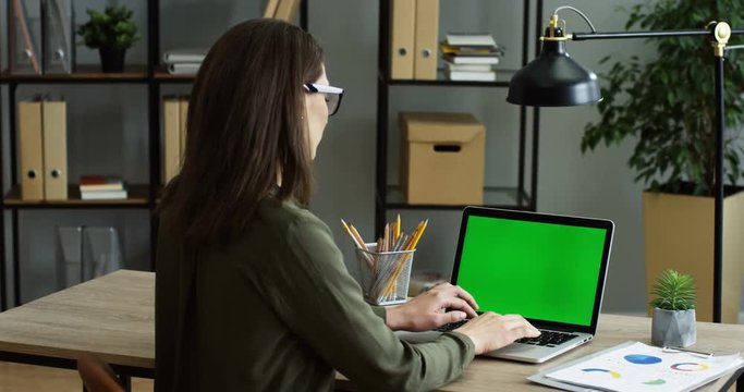 Rear of the woman sitting at the office desk, working while taping and texting on the keyboard of the laptop computer with green screen. Chroma key. Indoor