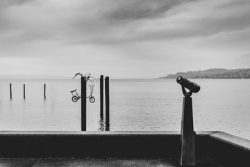 Black and white minimalist scene of a harbor with a coin-op viewer and several pillars with a...
