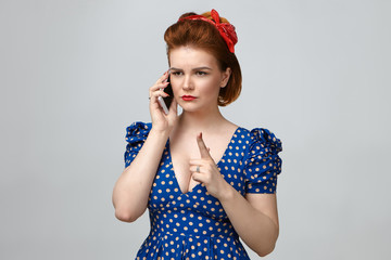 Picture of angry young mother in vintage outfit having strict look, raising index finger in front of her as warning sign, talking on smart phone with her naughty son who is still out with friends