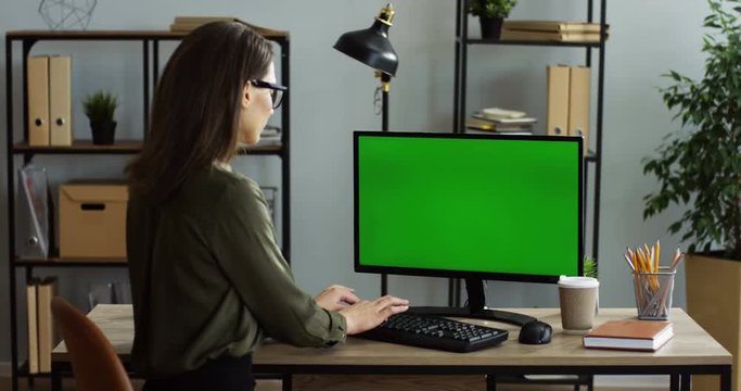 Rear of the caucasian young woman sitting at the table in the office and taping on the keyboard of the computer with green screen. Chroma key. Inside
