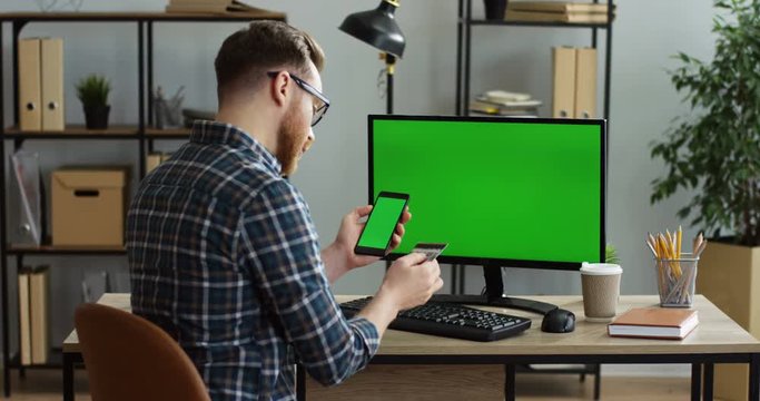 View from back on the young man in glasses sitting at the office table in front of the computer green screen and shopping online on the smartphone with a credit card in a hand. Chroma key. Indoors