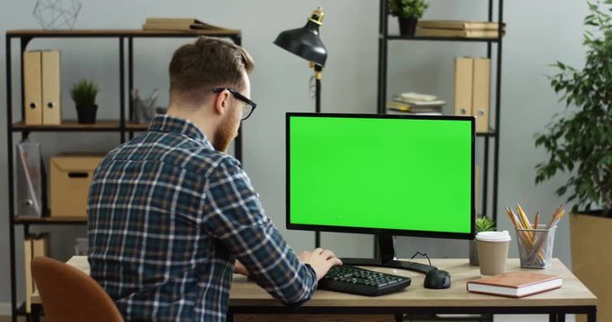 Rear of the caucasian young man sitting at the table in the office and taping on the keyboard of the computer with green screen. Chroma key. Indoor