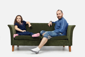 Horizontal shot of funny schoolgirl sitting on green sofa with her bearded father wearing casual clothes and staring at camera with emotional shocked expressions against white wall background © Anatoliy Karlyuk