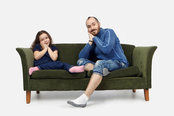 Bedtime. Picture of attractive positive young man in jeans and sweater sitting on sofa in living room, trying to send his daughter to bed, placing hands under cheek and smiling joyfully at camera