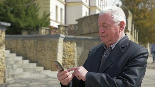 A 180 degree shot of a grey-headed man seeking business info on his smartphone in a  sunny street with a srone staircase in slow motion