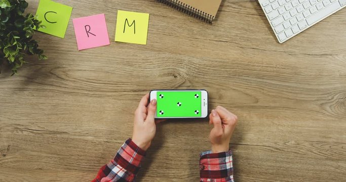Close up on the woman's hands taping on the white horizontal smartphone with a green screen on the wooden table. SRM. Chroma key. Tracking motion