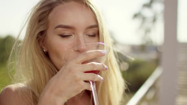 beautiful blonde brown-eyed with long hair girl smiling, showing off with her shoulder and sunglasses in brown swimsuit and bikini with wineglass of cocktail a background of nature