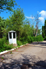 Fototapeta premium Metal arbor (watchman's house) with large glass windows in the edge of asphalt road, green trees park and blue cloudy sky background