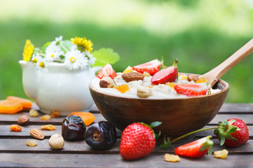 Oatmeal porridge in wooden bowl with fresh strawberries, mix nuts and dried fruit. Outdoor image, green meadow in background. Decorated with wild fowers in a small vase. 