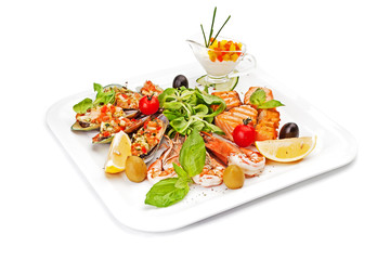 Pieces of fish fillet, fried and served on a white dish, served by leaves of salad, with olives, cherry tomatoes, an oiyster, arugula, a lemon segment, shrimps and tartlet with vegetables.