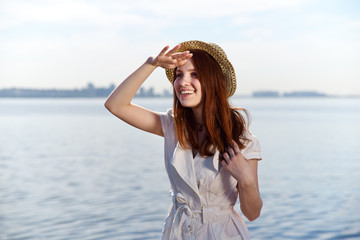 A beautiful smiling girl with red hair and a hat looks cheerfully into the distance, putting her palm to her forehead.