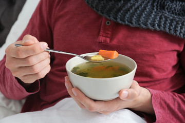 Sick young man eating broth to cure cold in bed at home