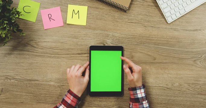 Close up on the woman's hands taping on the black vertical tablet device with a green screen on the wooden table. SRM. Chroma key.