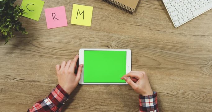 Close up on the woman's hands taping on the white horizontal tablet device with a green screen on the wooden table. SRM. Chroma key.