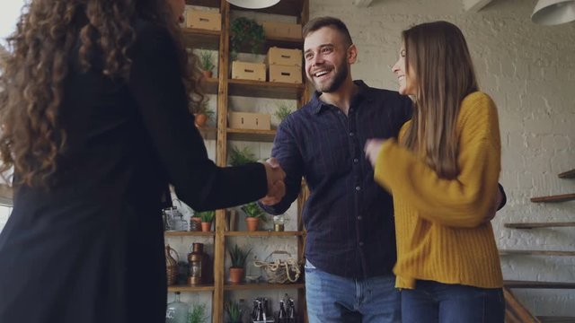 Happy young couple is buying new house shaking hands with female realtor then hugging and laughing. Relocation, happiness and accommodation concept.
