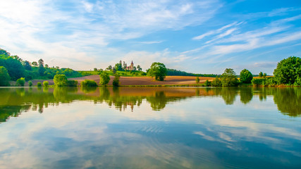Fototapeta na wymiar Romantic landscape with small church on the hill reflected in the pond. Sunny summer day with blue sky and white clouds. St. Peter and Pauls church at Bysicky near Lazne Belohrad, Czech Republic.
