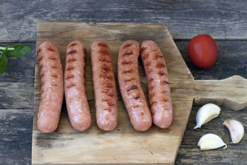Grilled sausages located on a cutting board which is located on an old wooden background in a rustic style, tomatoes, garlic and greens