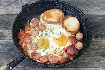 Breakfast in a rustic style, fried eggs with sausages tomatoes greens and croutons in an old frying pan on a wooden background