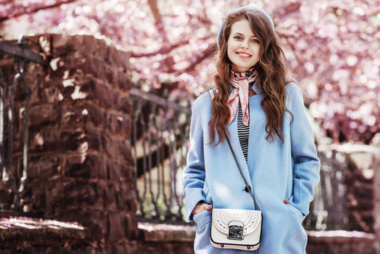 Outdoor portrait of young beautiful fashionable happy smiling girl wearing stylish pastel blue coat, beret, scarf, with small white perforated bag. Spring fashion concept. Copy, empty space for text