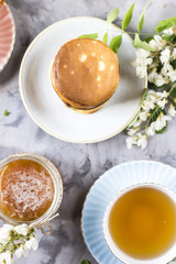 A pile of fritters next to a cup of tea and a jar of honey on a gray background among the flowers of the acacia. Spring delicious breakfast. Top view, flat lay