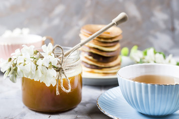 A pile of fritters next to a cup of tea and a jar of honey on a gray background among the flowers of the acacia. Spring delicious breakfast