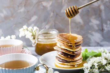 A pile of fritters next to a cup of tea and a jar of honey on a gray background among the flowers of the acacia. Spring delicious breakfast