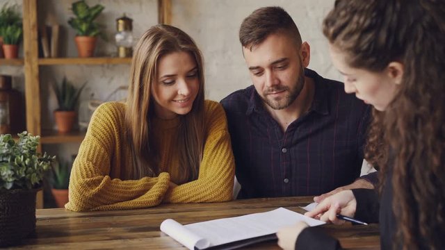 Female realtor is reading purchase and sell agreement to young couple, man is signing document and taking key, shaking hands with agent and kissing his wife.