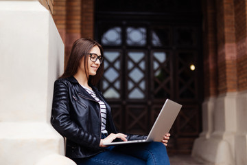 Fototapeta na wymiar Young beautiful brunette student in glasses sits on bench at university and works on laptop.