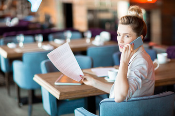 Young busy woman with smartphone and financial papers sitting in armchair in modern cafe or restaurant