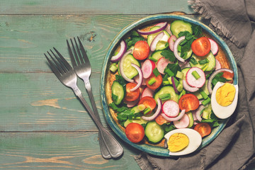 Vegetable salad with boiled egg served on a plate. Green rustic background, forks, napkin. Top view, copy space. Toned photo