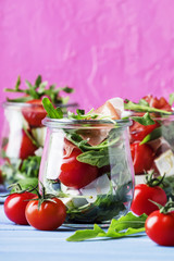 Salad with arugula, cheese, cherry tomatoes and prosciutto in glass jars, pink blue background, selective focus