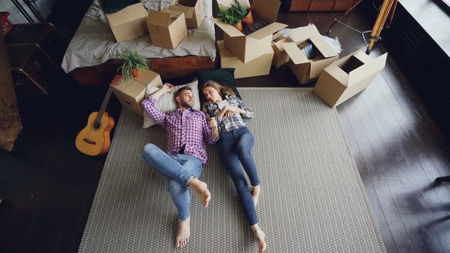 Top view of loving couple lying on bedroom floor in new apartment and talking. Guitar, packed things in carton boxes, double bed and modern carpet are visible.