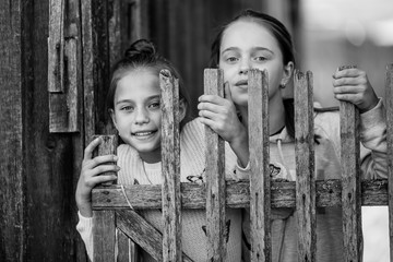 Portrait of two sisters girls teenagers look out from behind a wooden village fence. Black and white photo.