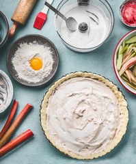 Rhubarb cake preparation. Tart baking pan with pie dough and cream fresh on kitchen table background with rolling pin, flour, egg and fresh red rhubarb stalks, top view. Seasonal cooking concept
