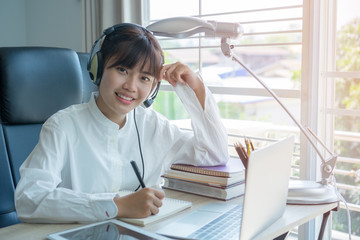 Student learning online study concept: Beautiful Asian girl listening with Headphones and laptop, sitting smile writing notes in textbook at her desk in home for e-learning in educational technology